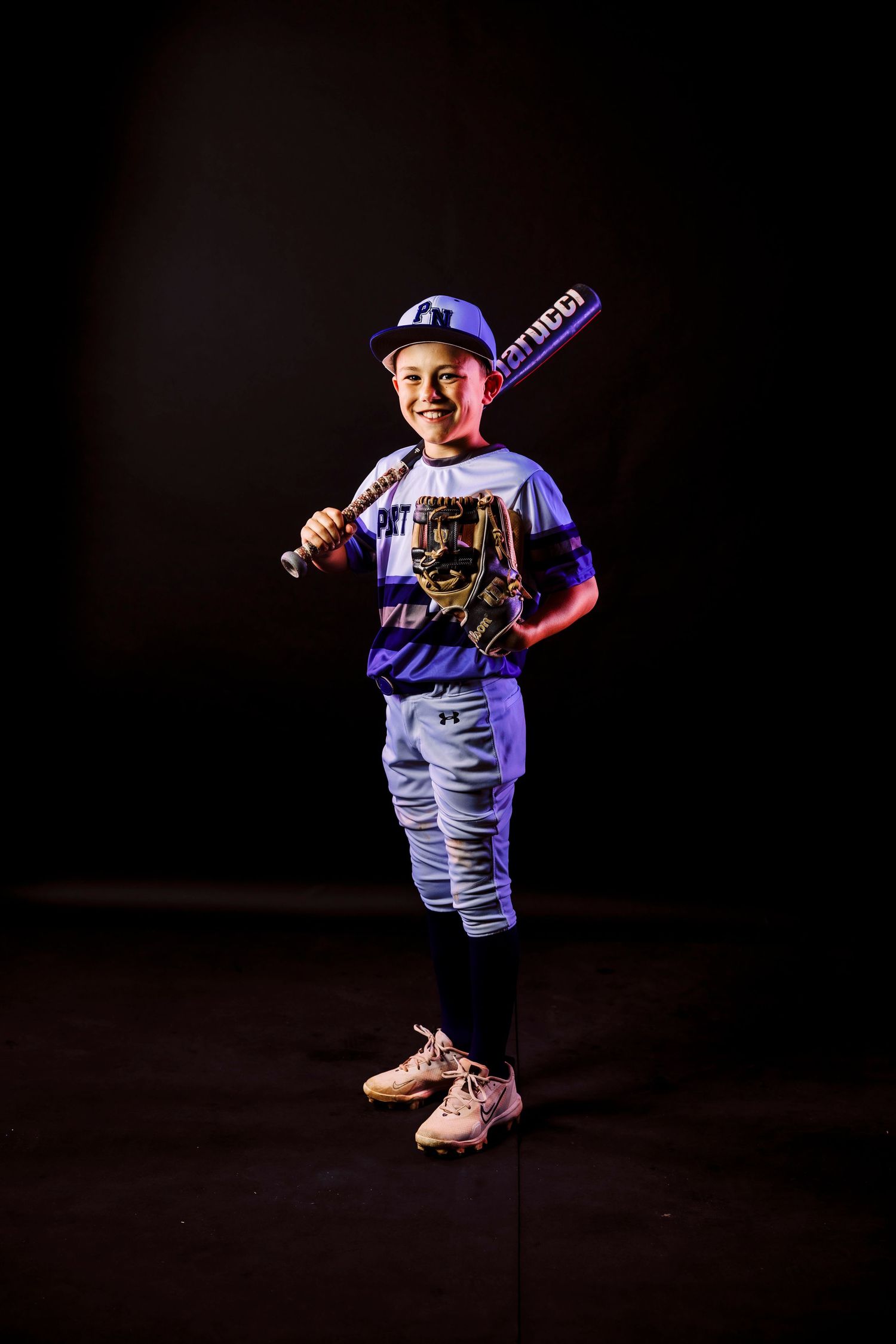 Young baseball player in uniform poses with bat against dark background with dramatic lighting.