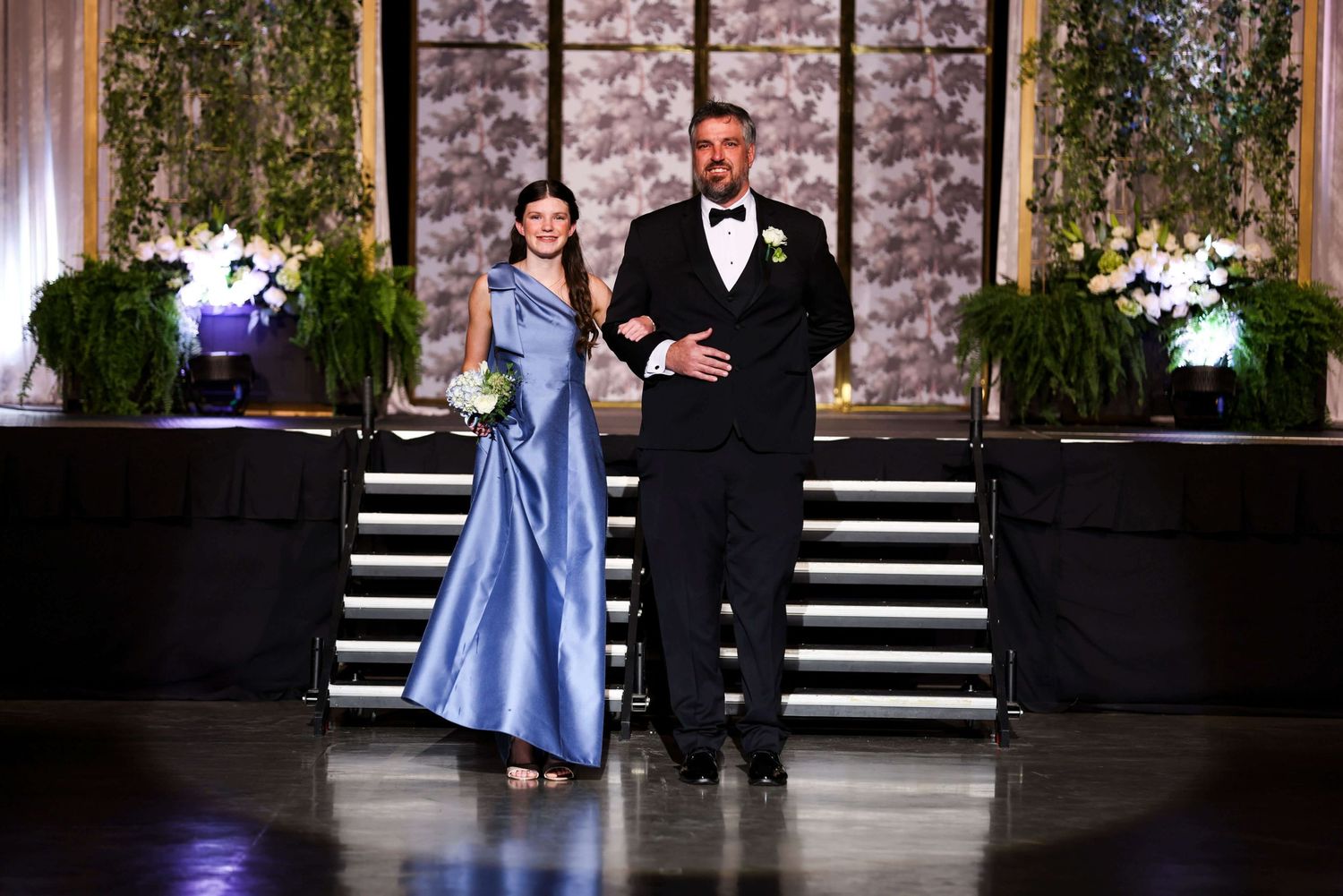 Couple in formal attire walk down stone steps during an elegant outdoor event.