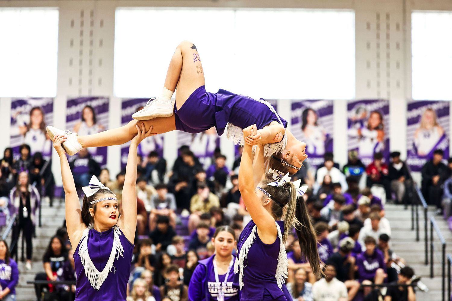 Cheerleaders performing aerial stunt with purple uniforms at indoor competition.