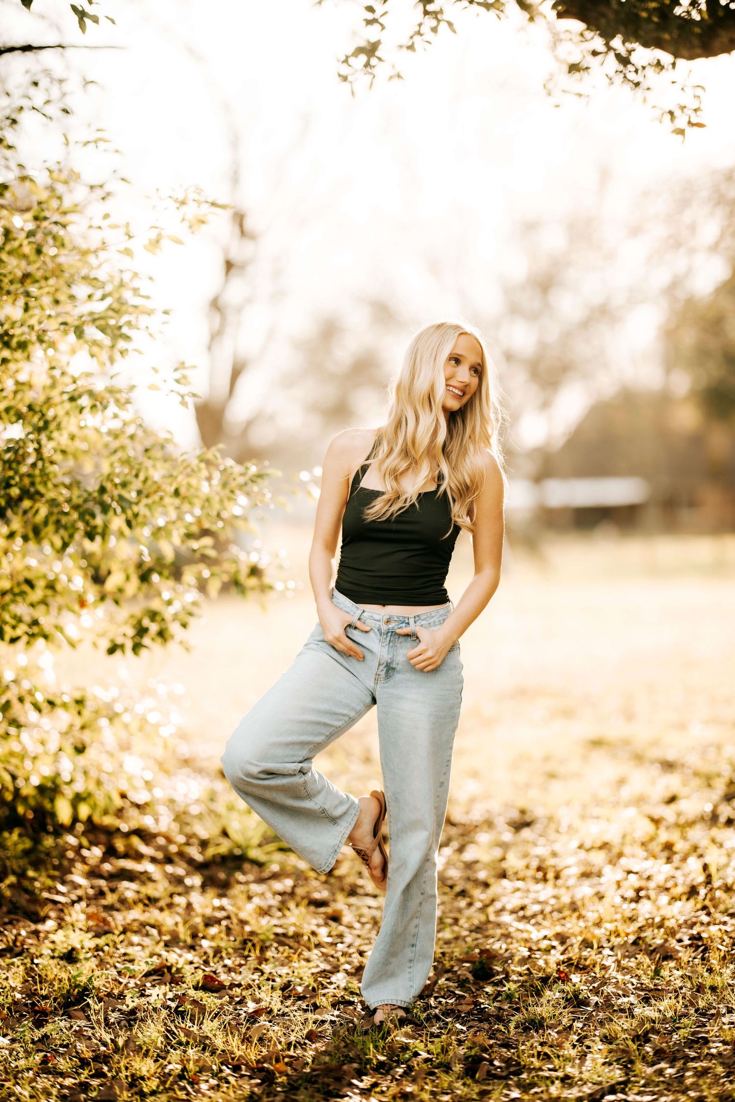 A person in casual attire poses against a sunlit autumn background with fallen leaves.