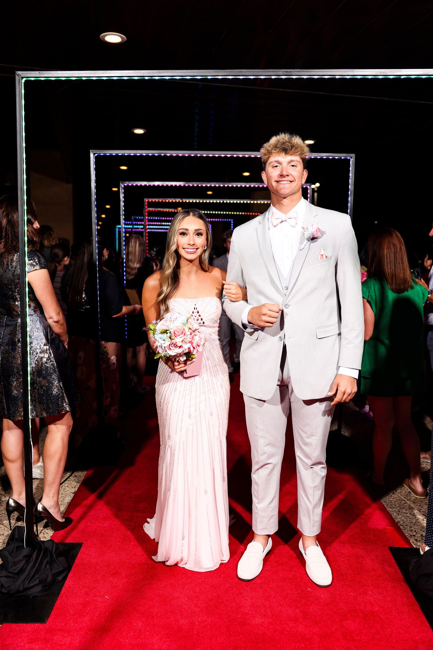 A couple poses on a red carpet at a formal event wearing coordinating white and pink formal attire.