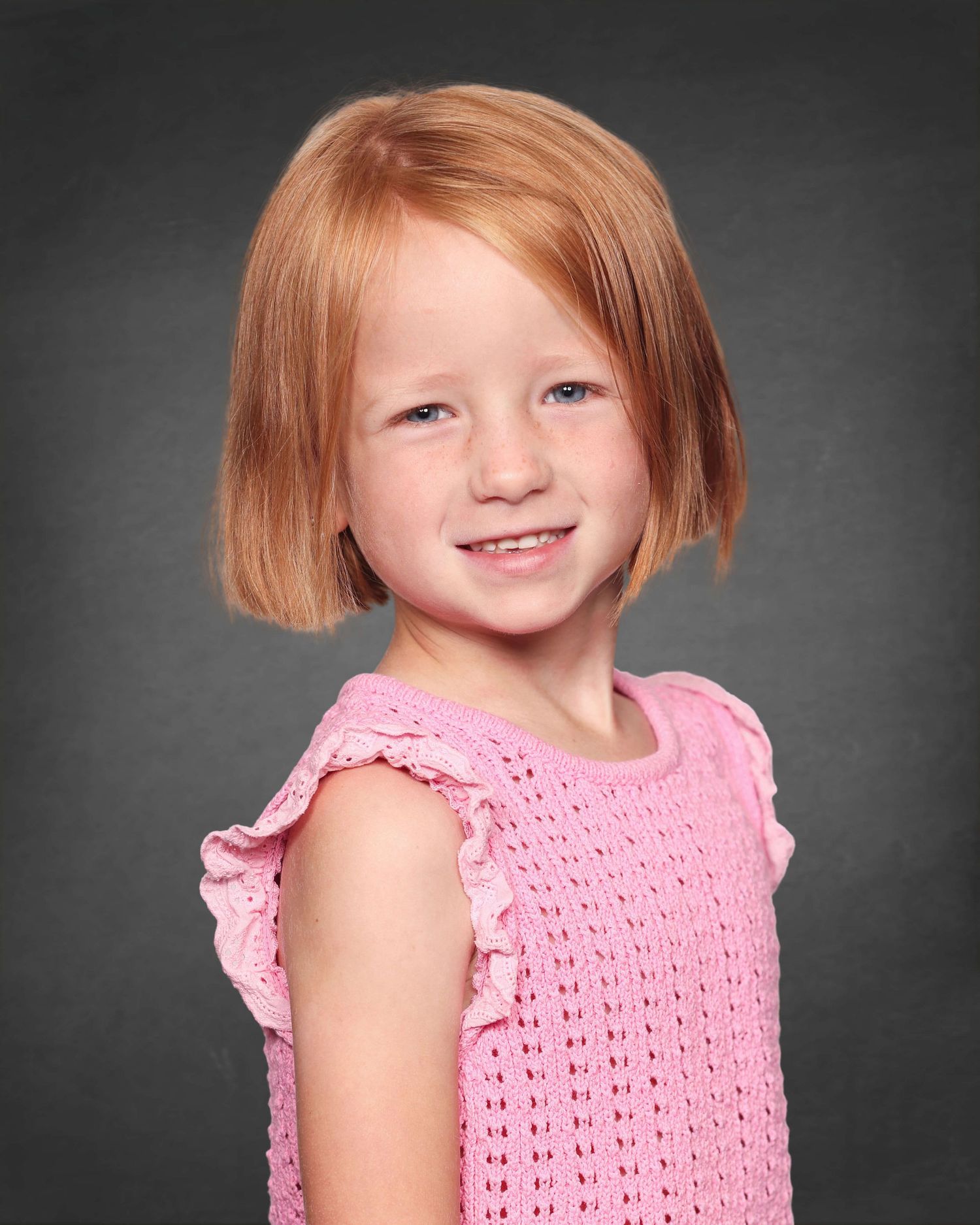 Studio portrait of a young child with strawberry blonde hair wearing a pink knit top against a dark gray background.