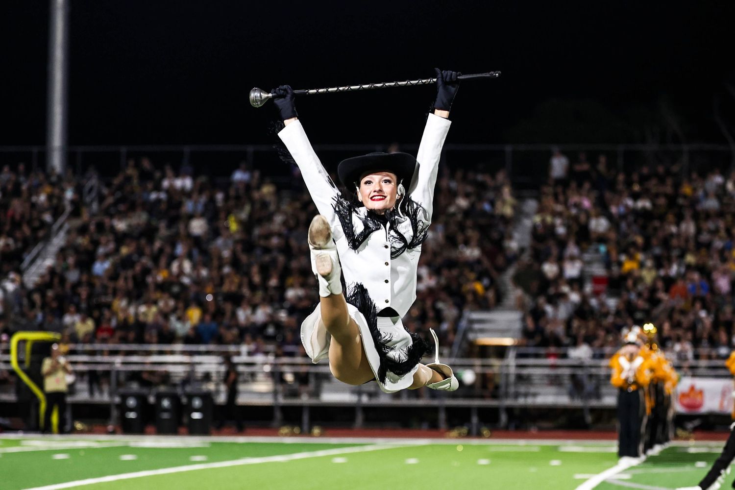 Marching band performer in black uniform executing aerial baton toss at night.