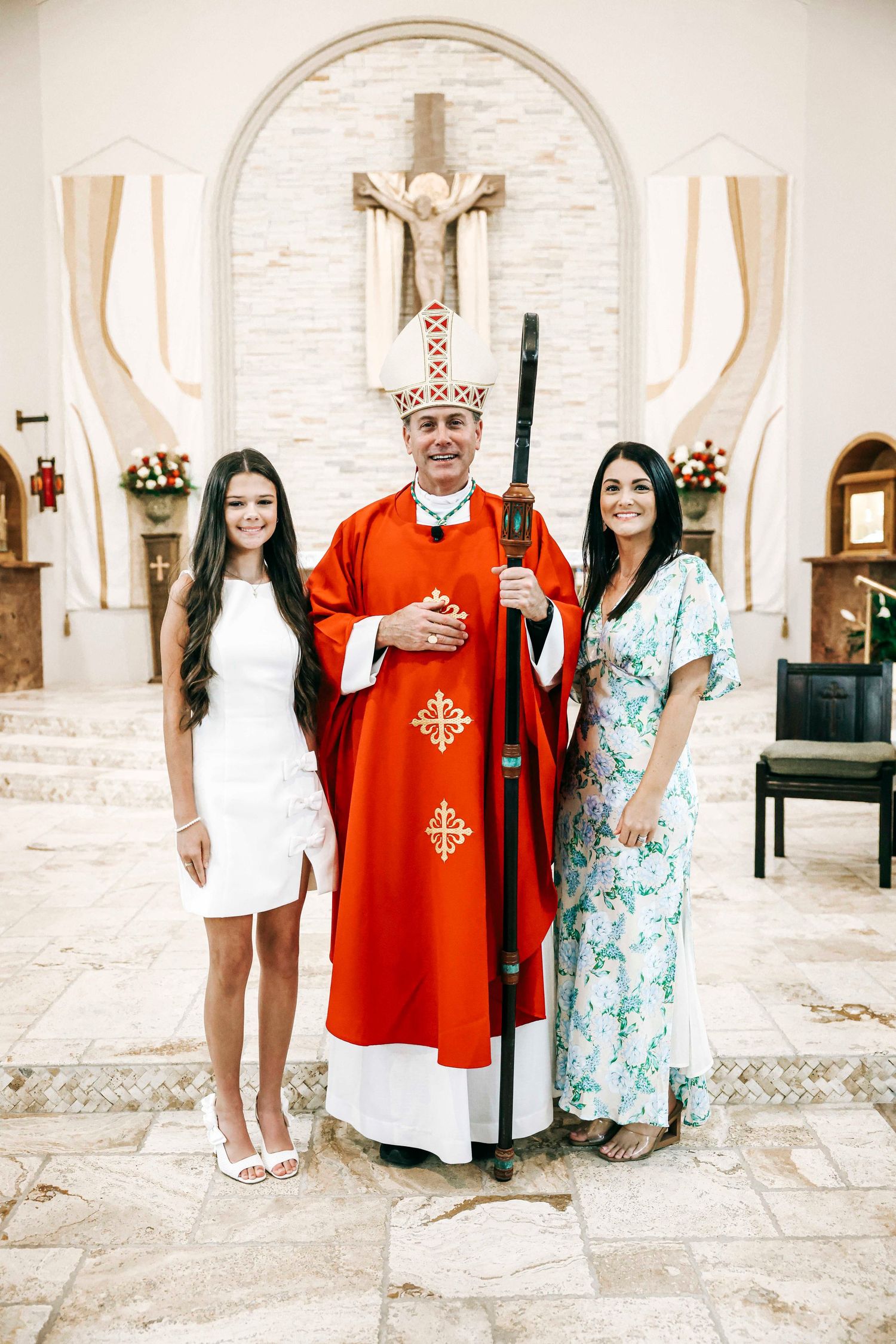 A Catholic bishop in traditional red vestments stands in a church sanctuary with a crucifix visible on the wall behind.