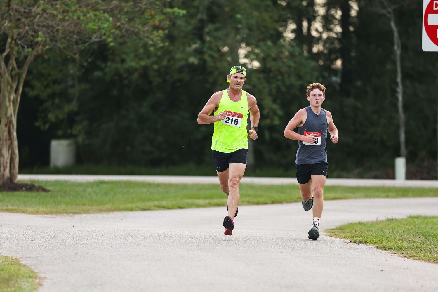 Two runners in athletic gear compete on a paved path through a park during a daytime race.