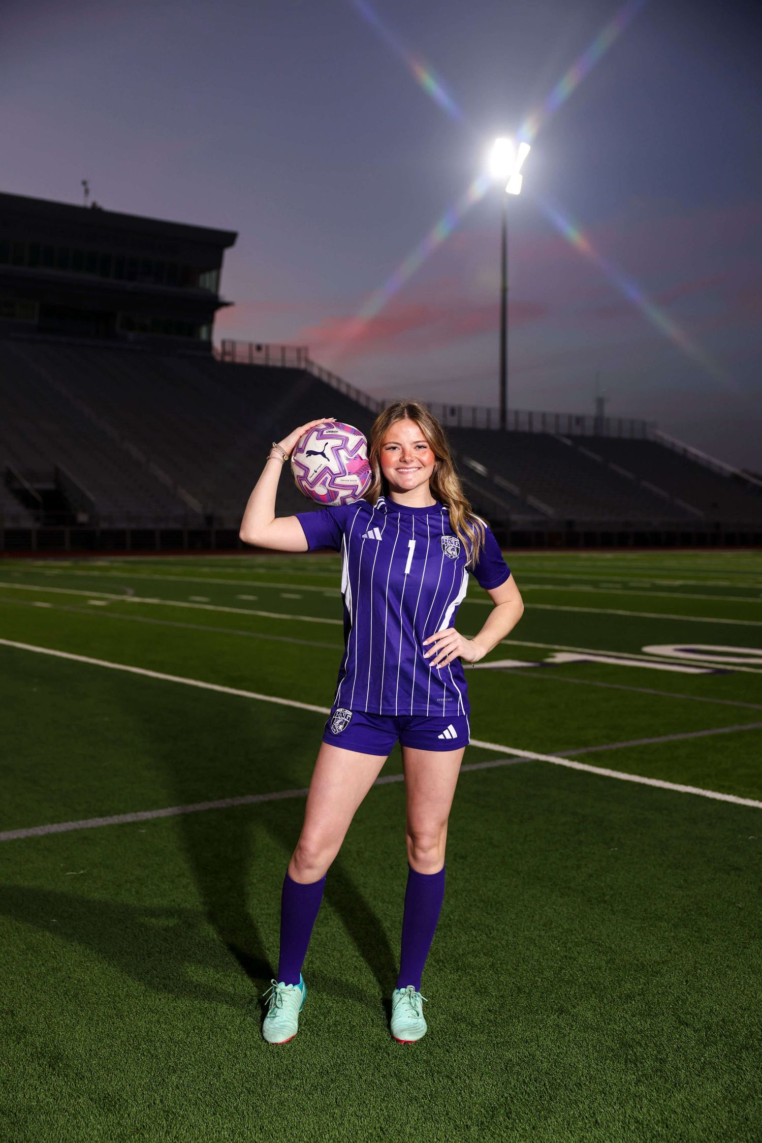 Soccer player poses with ball on field at night under stadium lights.