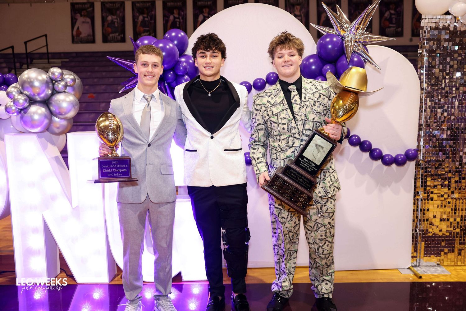 Students in formal attire gather at a school dance with purple and silver balloon decorations and festive lighting.