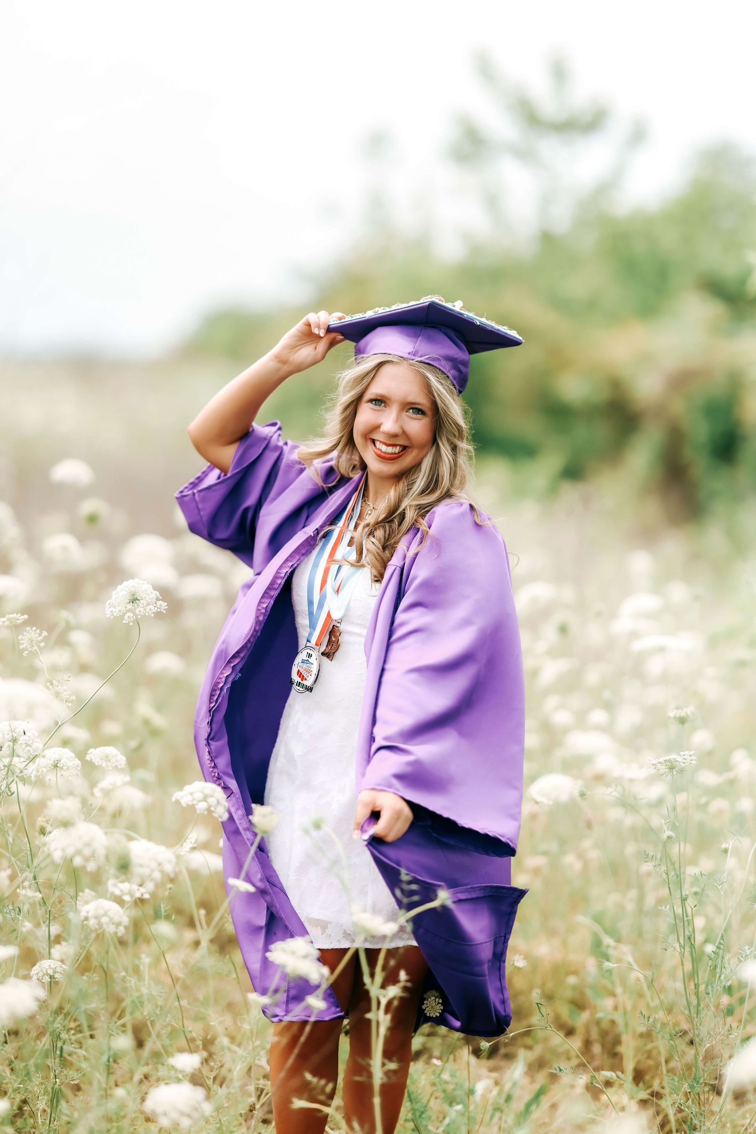 Graduation photoshoot in purple cap and gown amid white wildflowers.