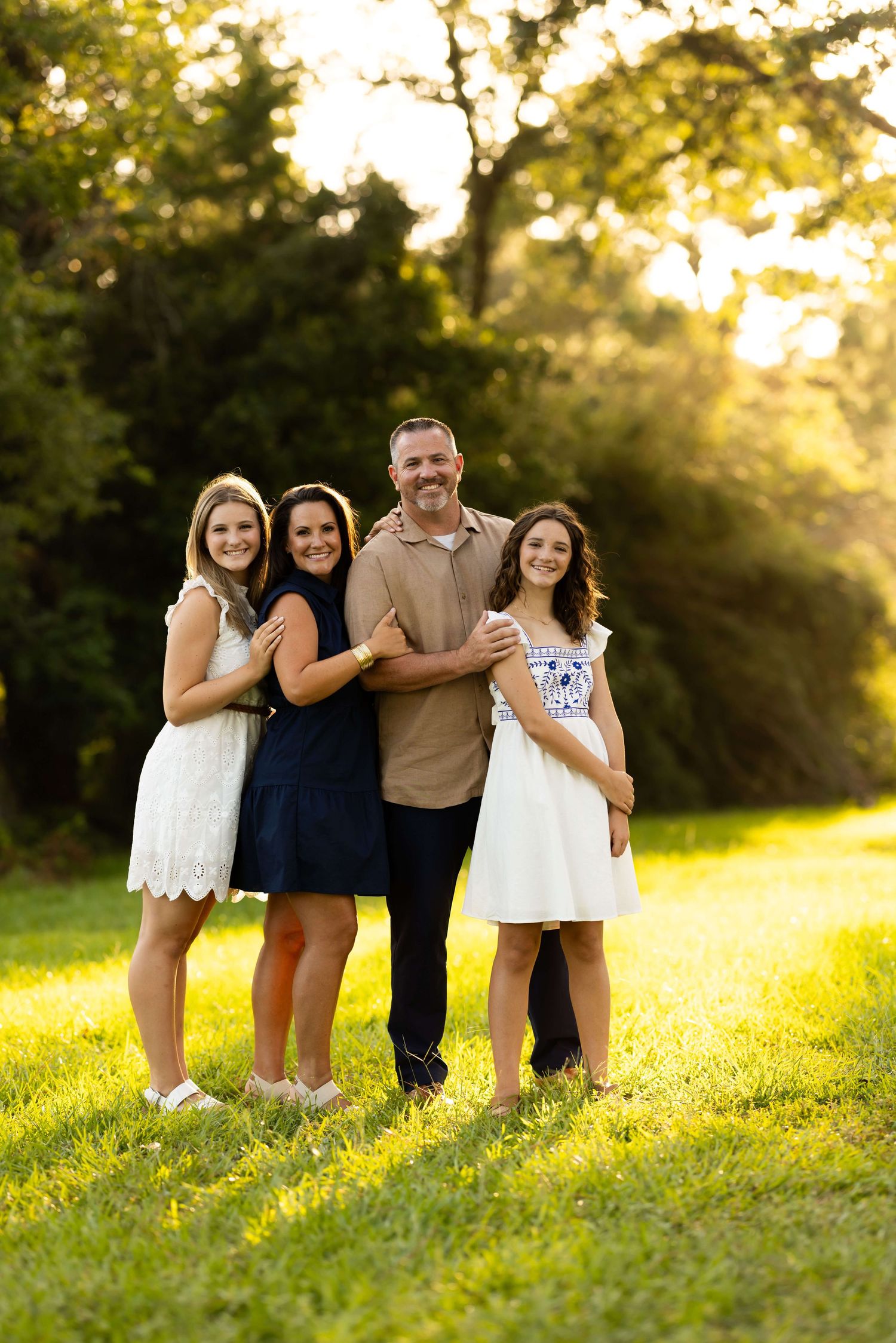 Family of five posing together on grass during golden hour with sunbeams streaming through trees.