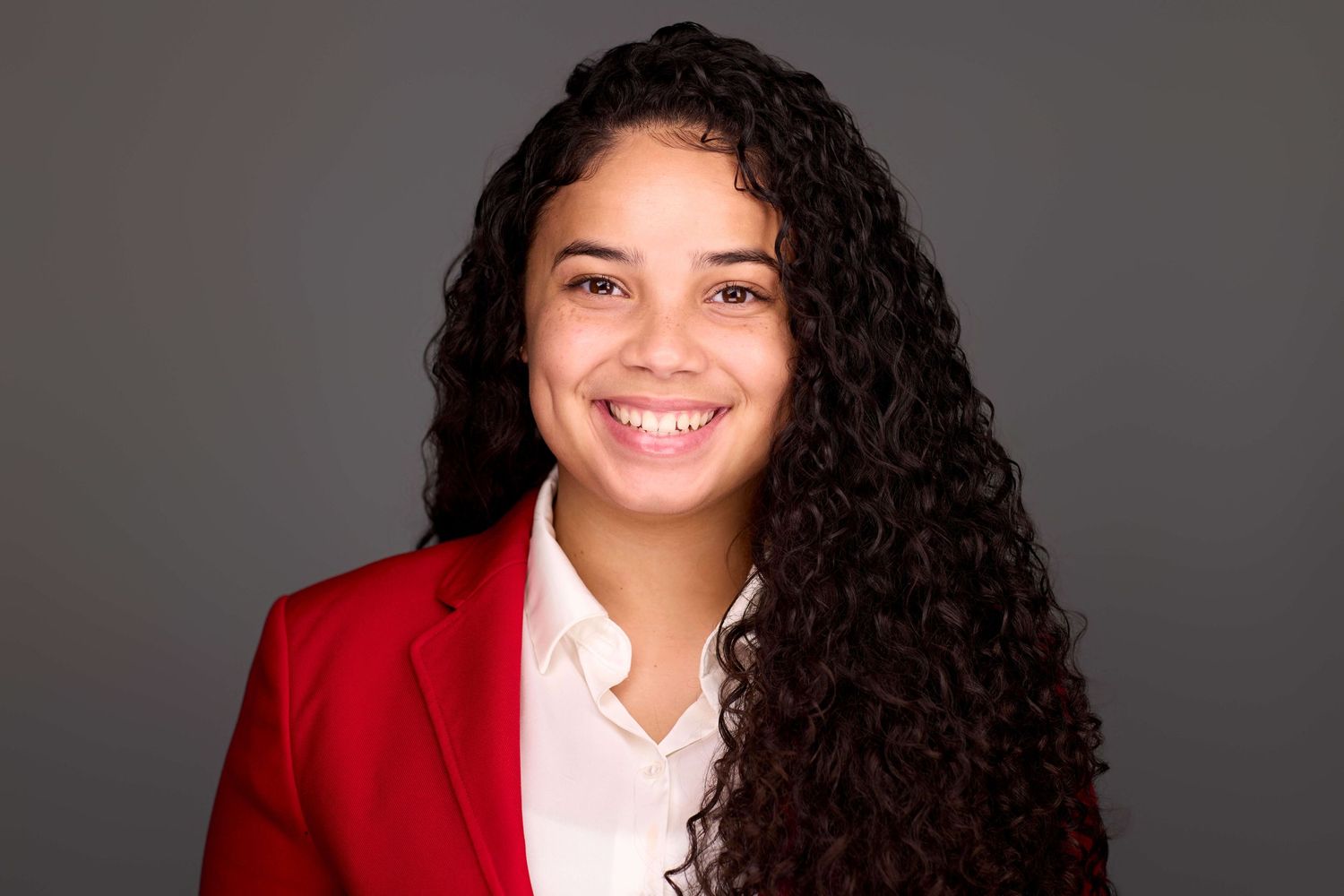 Professional portrait of person in red blazer with curly dark hair against gray background.