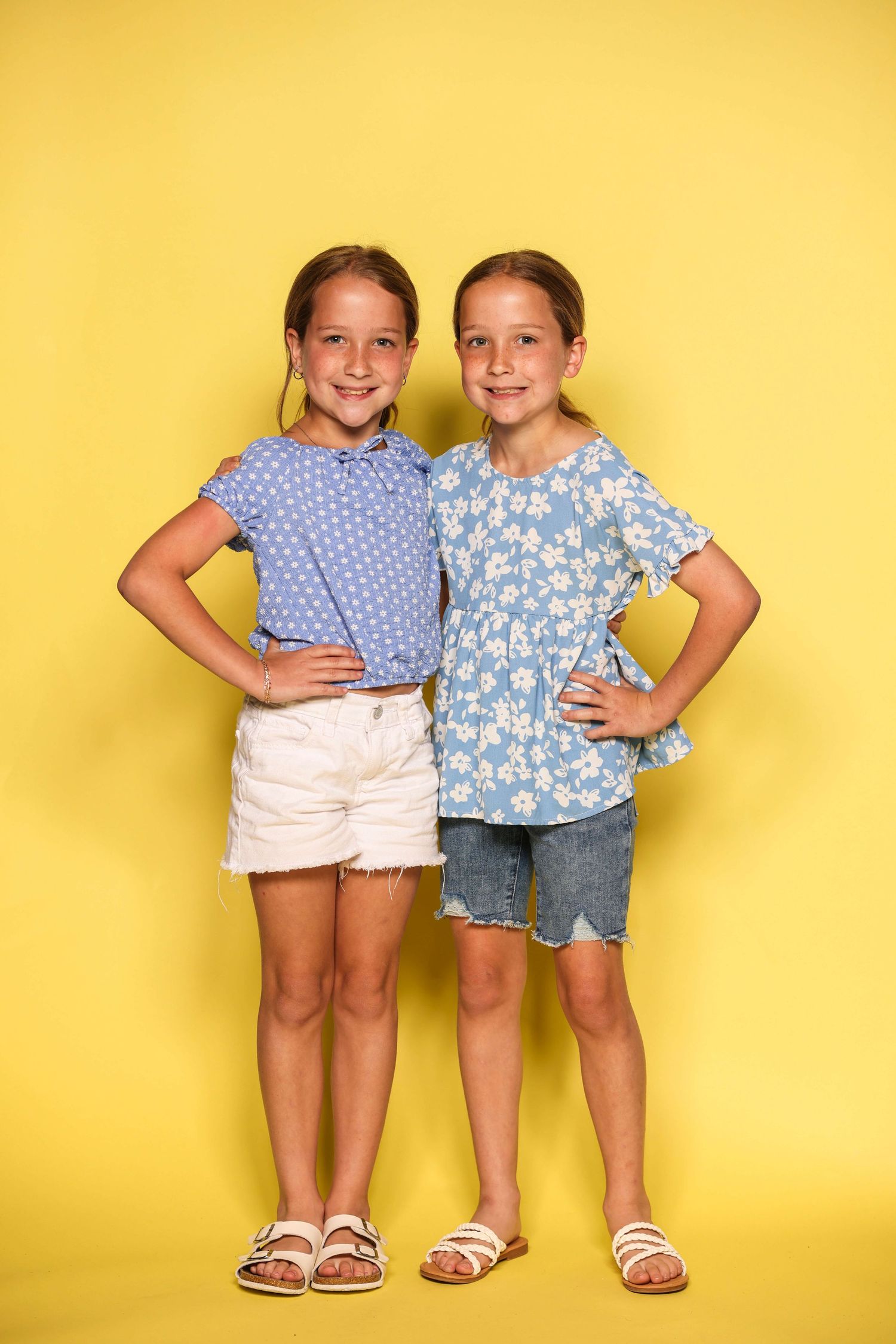 Two young girls in summer outfits pose with hands on hips against a bright yellow background.