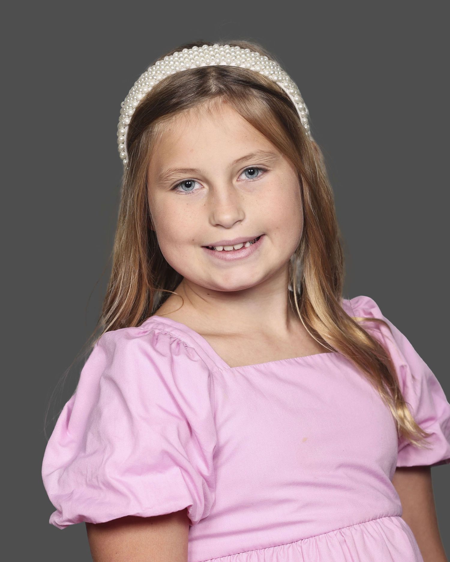 Young child wearing a pink puff sleeve shirt and sparkly headband smiles at the camera against a gray background.