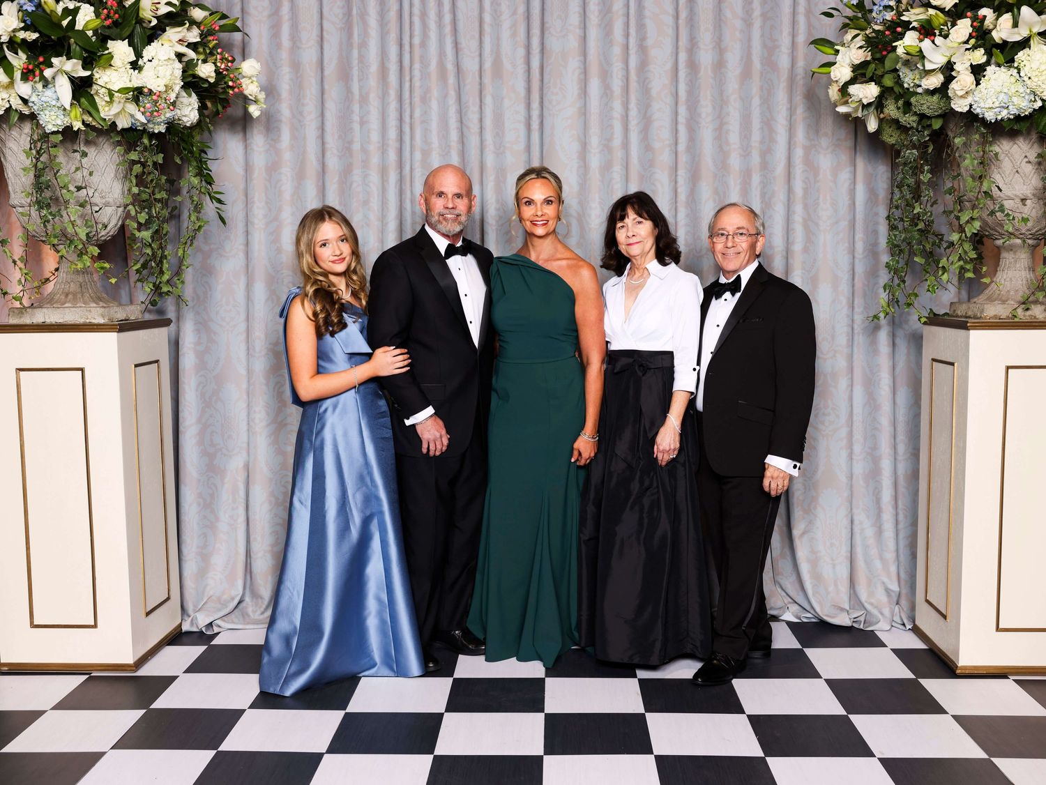 Wedding party poses for formal photo against grey curtain backdrop with white floral arrangements.