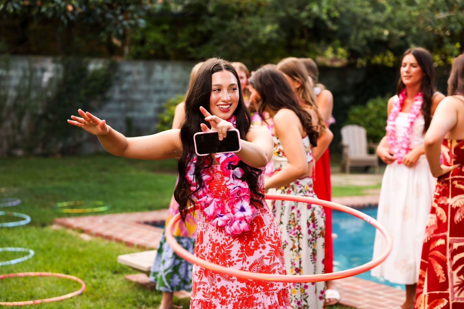 A group of people in floral dresses playing outdoor party games with hula hoops near a swimming pool.