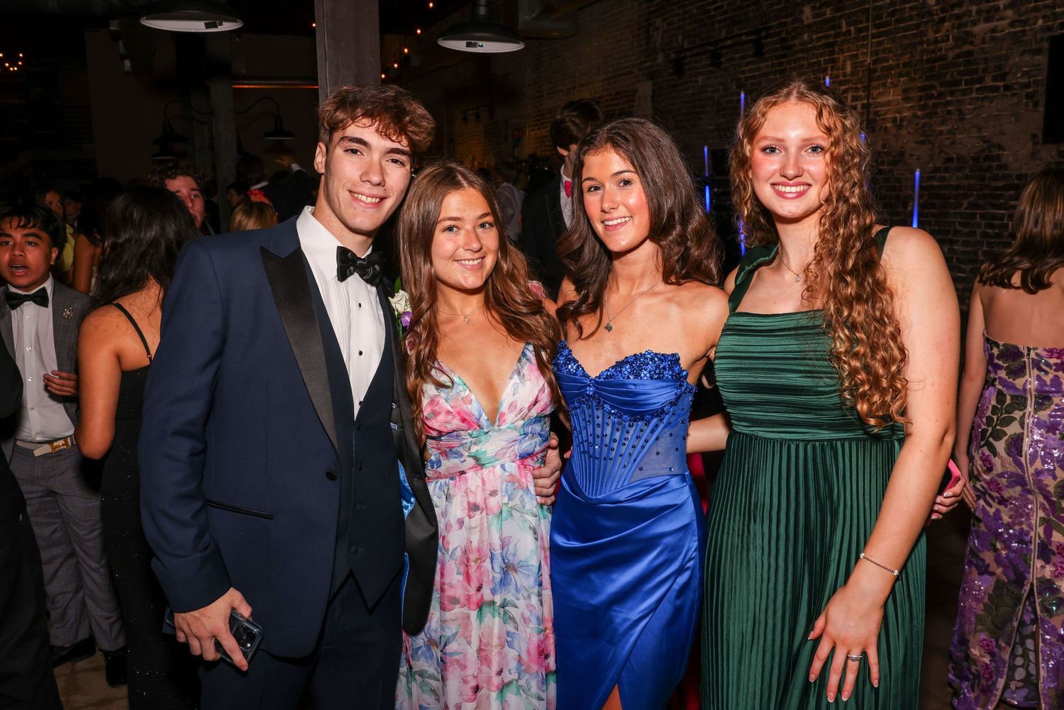 Four students dressed in formal attire smile together at a prom event in front of exposed brick walls.