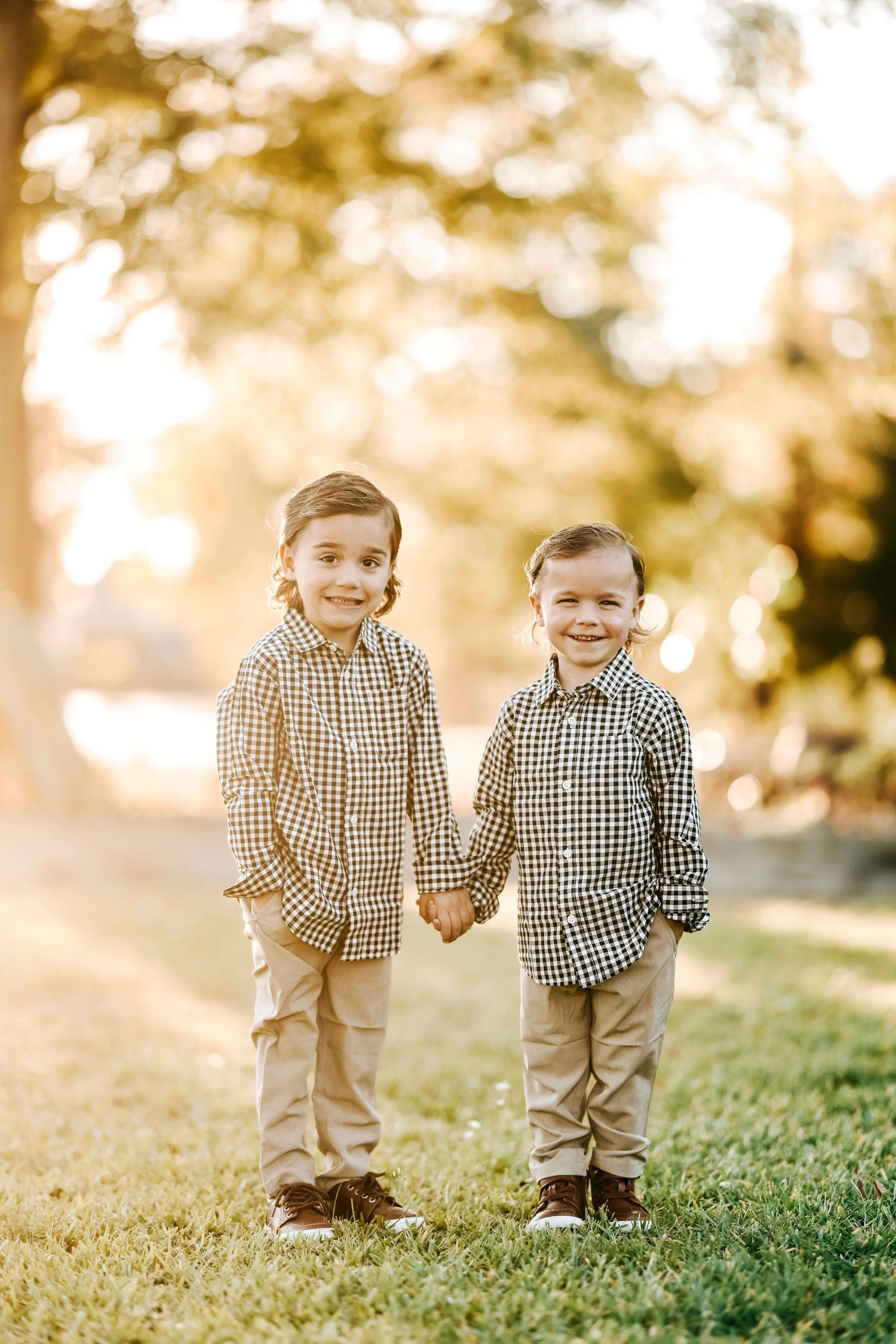 Two young children in matching plaid shirts and khaki pants holding hands outdoors at golden hour.