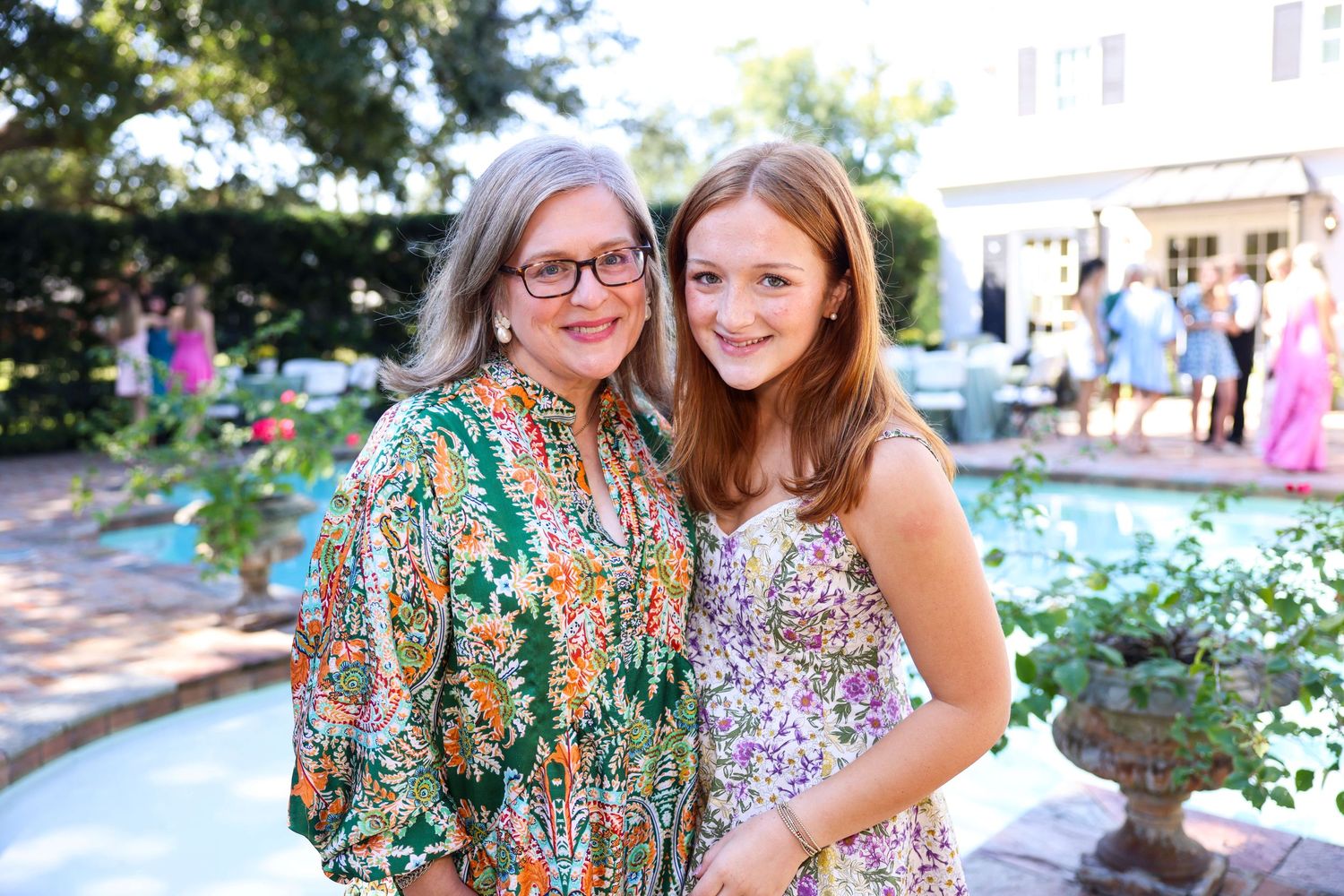 Two people in colorful summer outfits smile together outdoors near flowering plants and greenery.