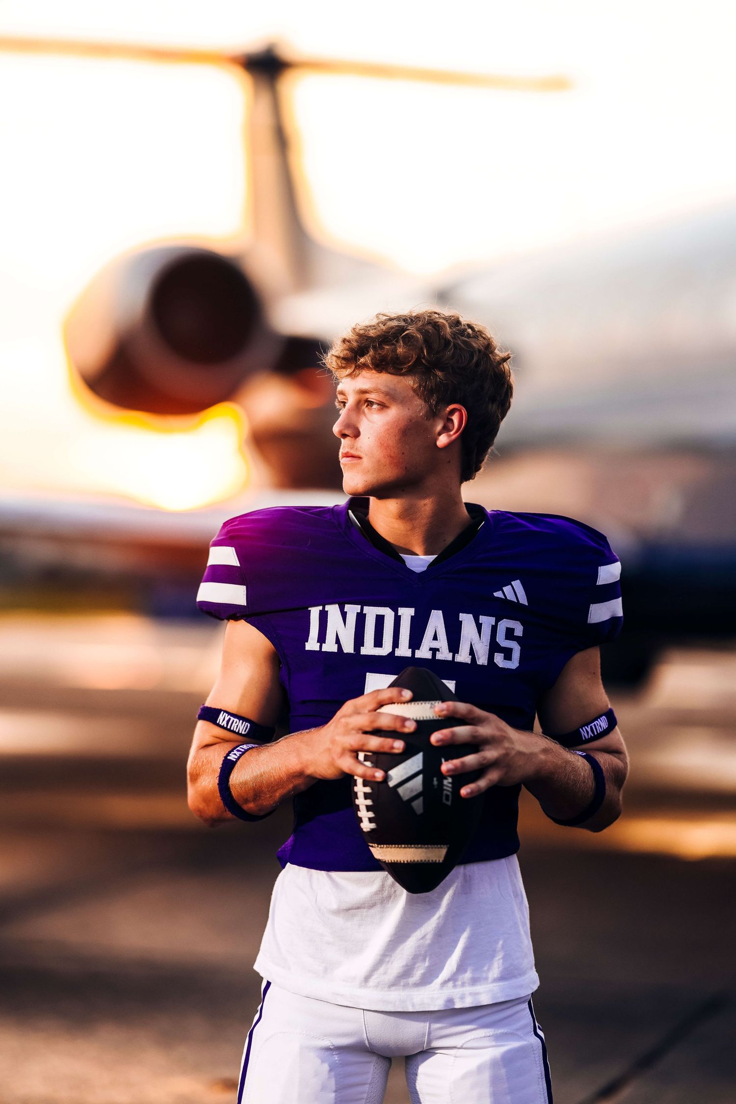 Football player in purple Indians jersey holds ball against dramatic sunset backdrop.