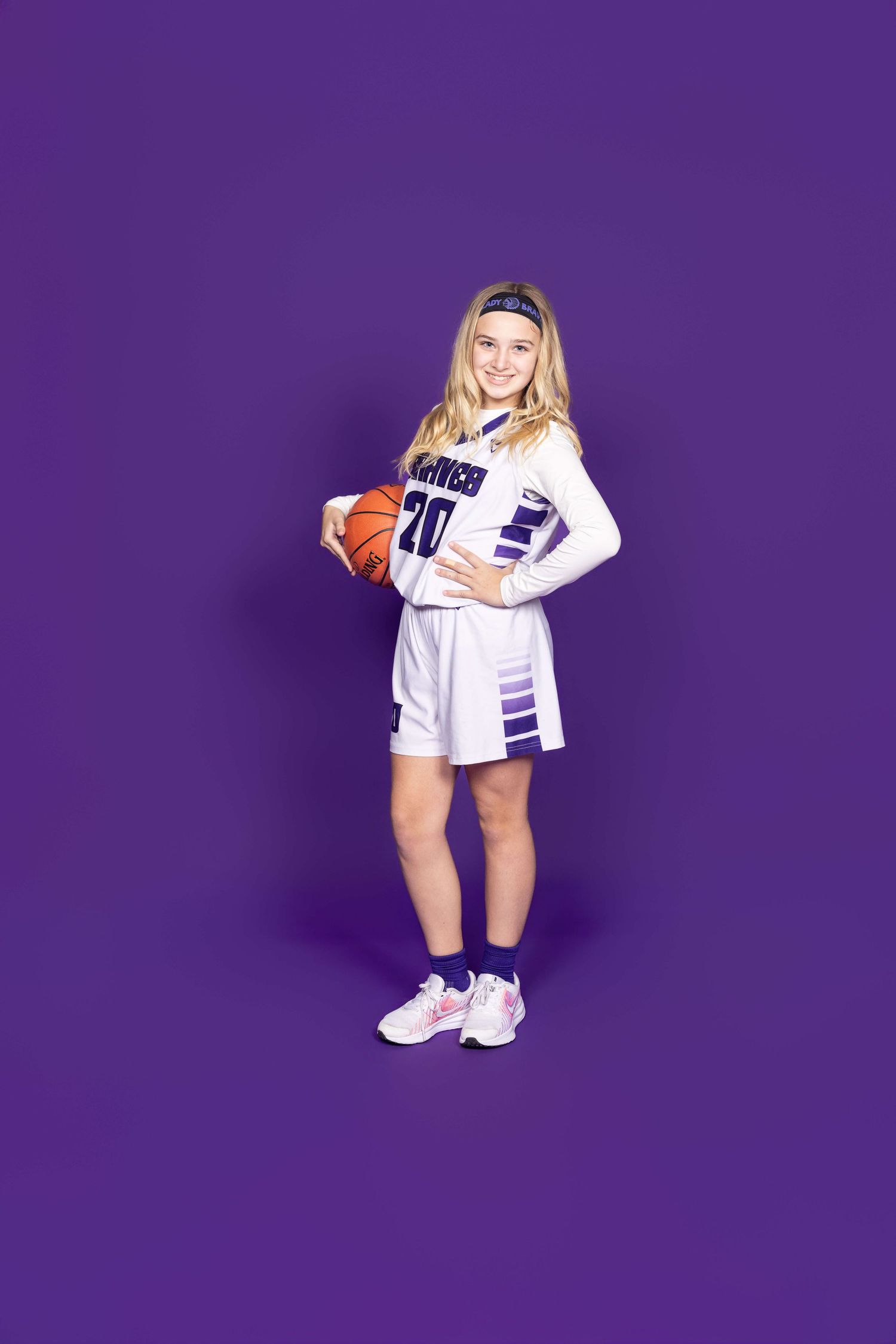 Basketball player in white and purple uniform poses with ball against purple backdrop.