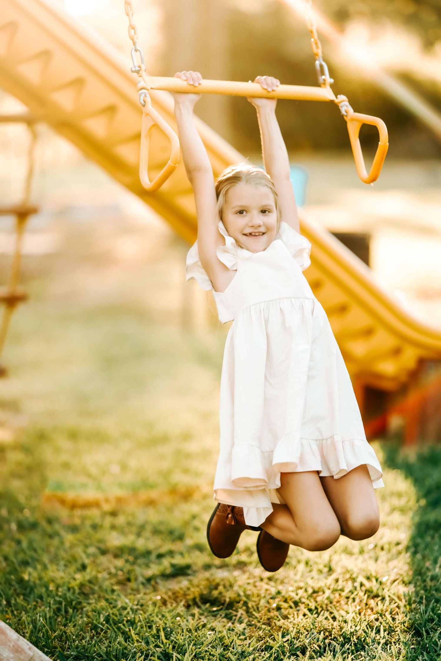 Child in white dress joyfully swinging on playground equipment during golden hour.
