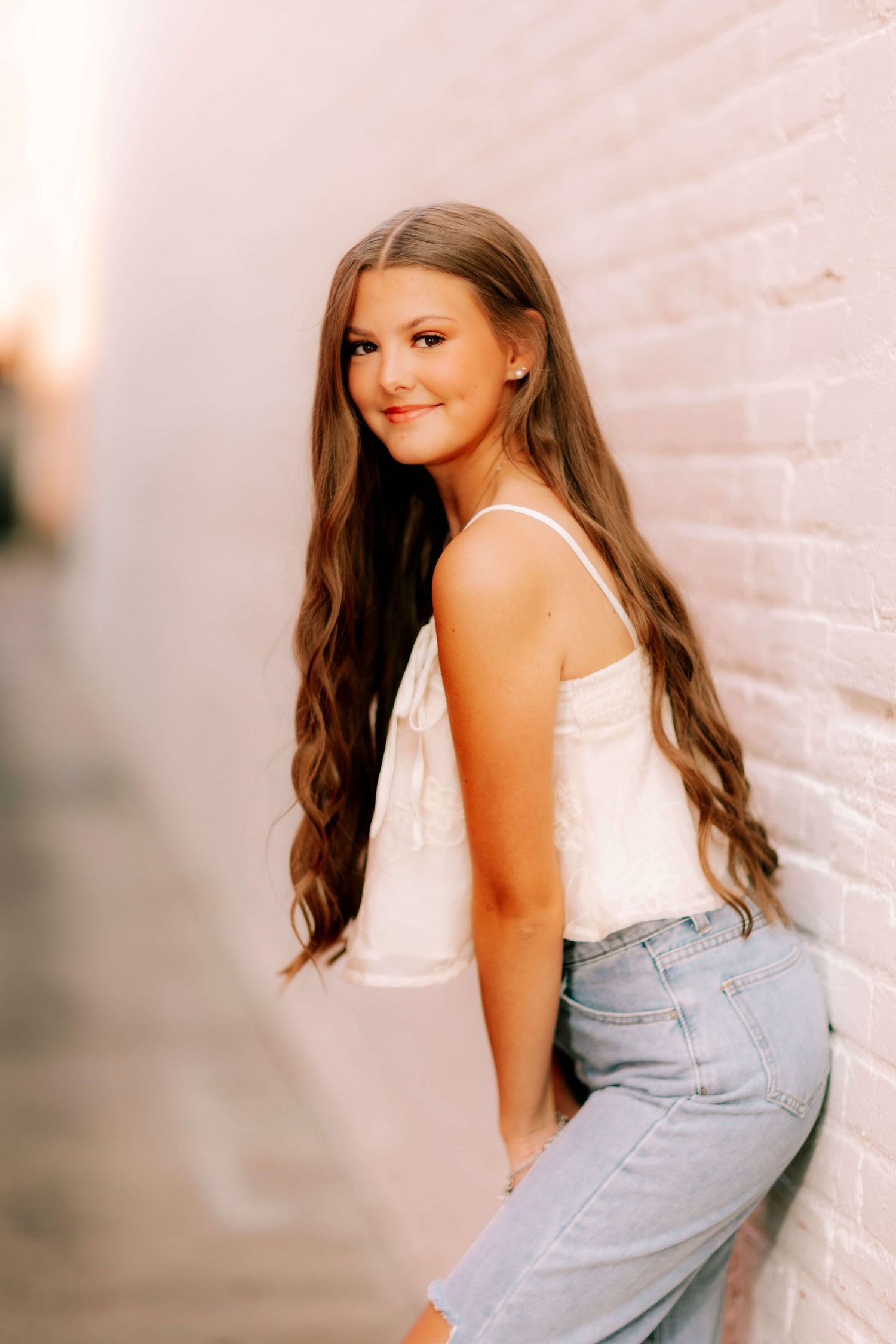 Woman in white top and jeans poses playfully against light colored wall.