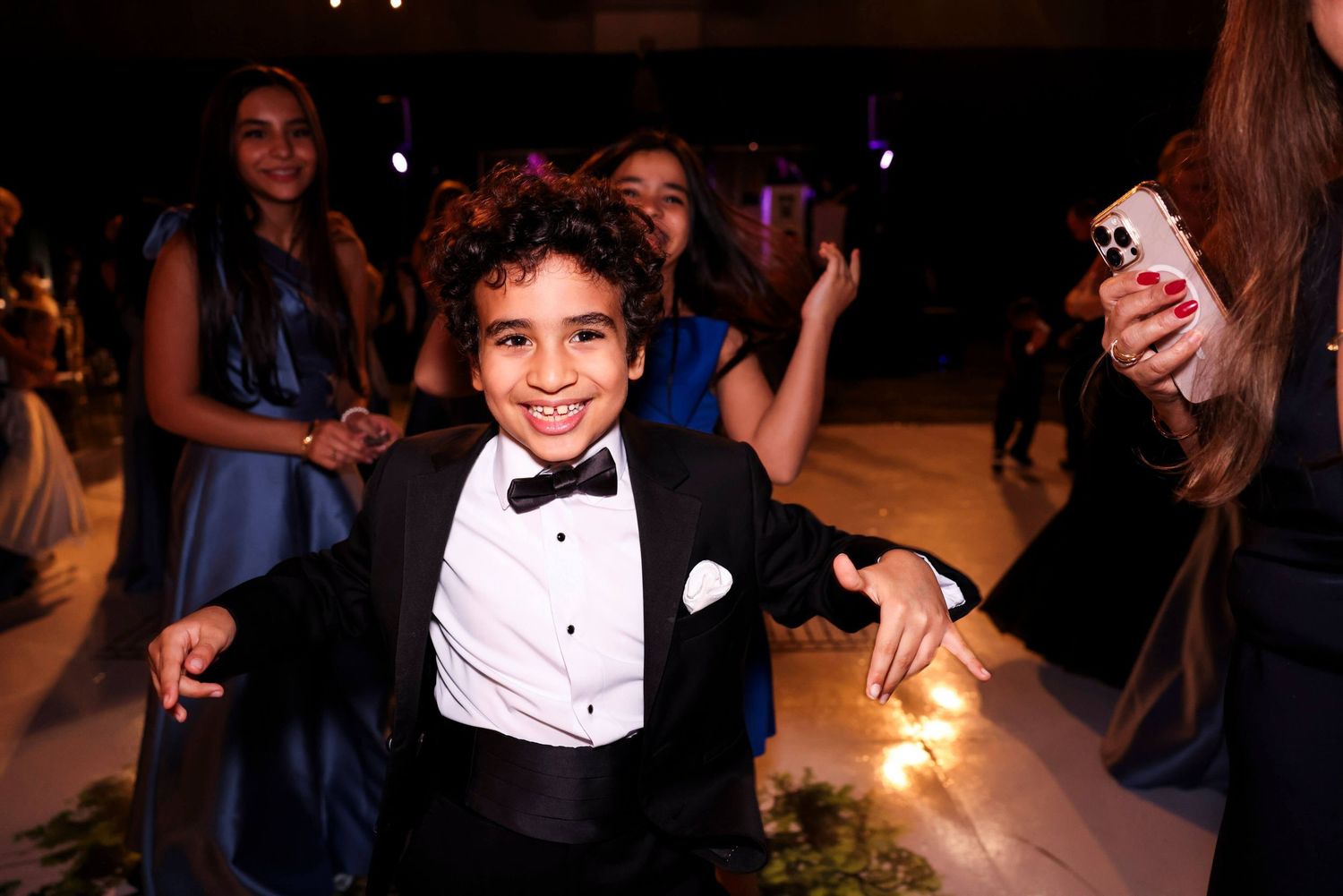 Young guest in a tuxedo and bowtie smiles brightly at a formal evening event with dancing in the background.