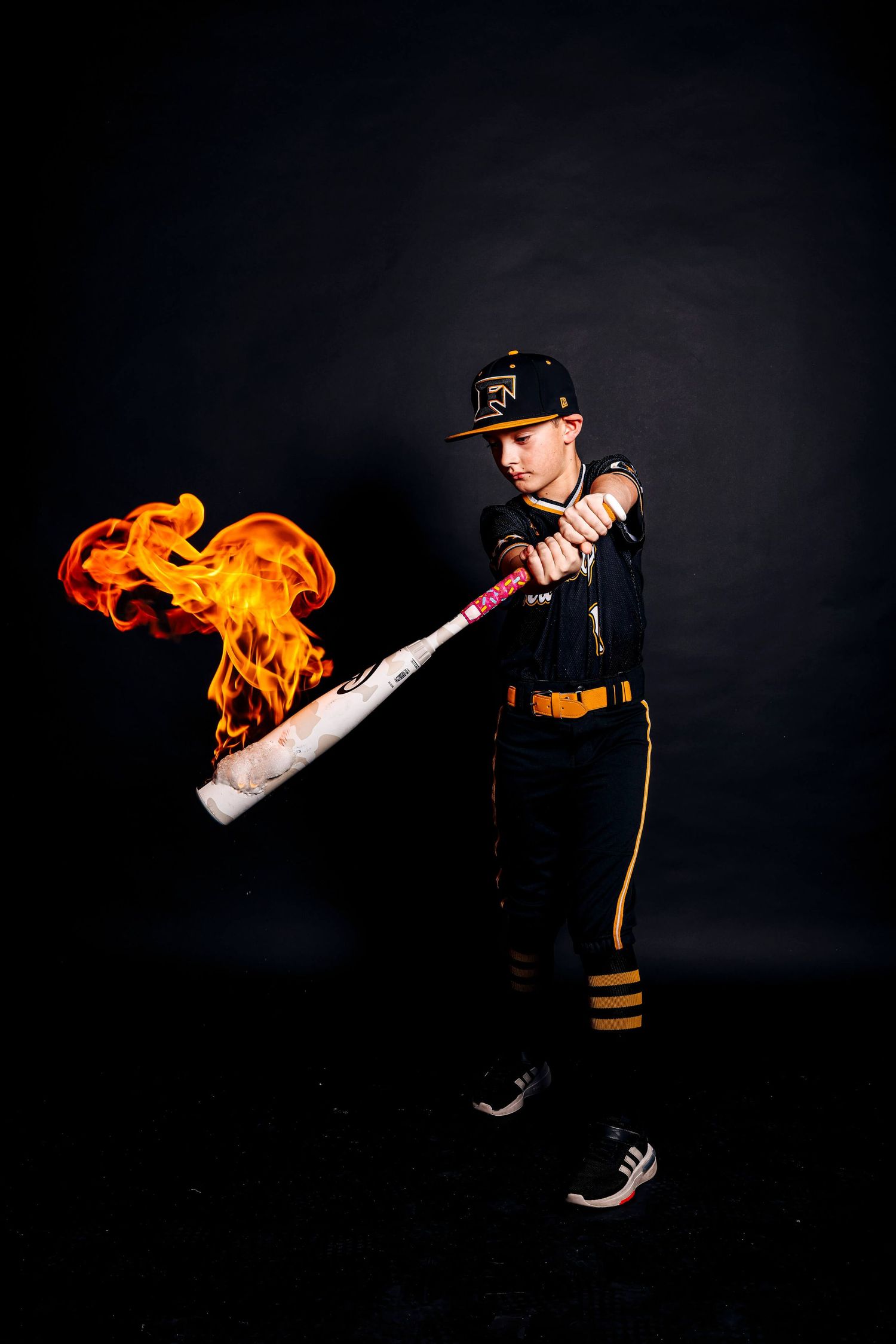 Baseball player in black uniform swings flaming bat against dark background in dramatic action shot.