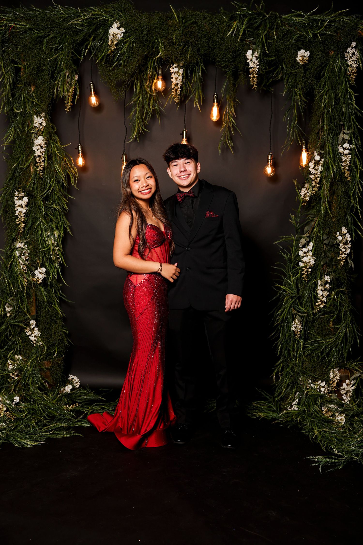 A couple poses in formal attire under an arch decorated with greenery, white flowers and hanging Edison bulbs.