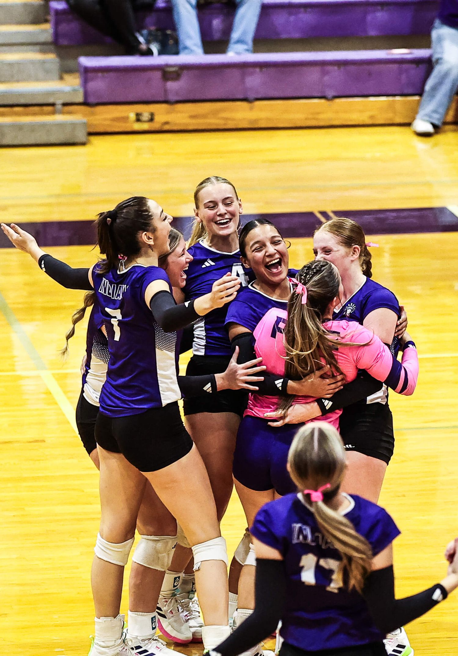 Volleyball team celebrates victory on indoor court wearing purple and black uniforms.