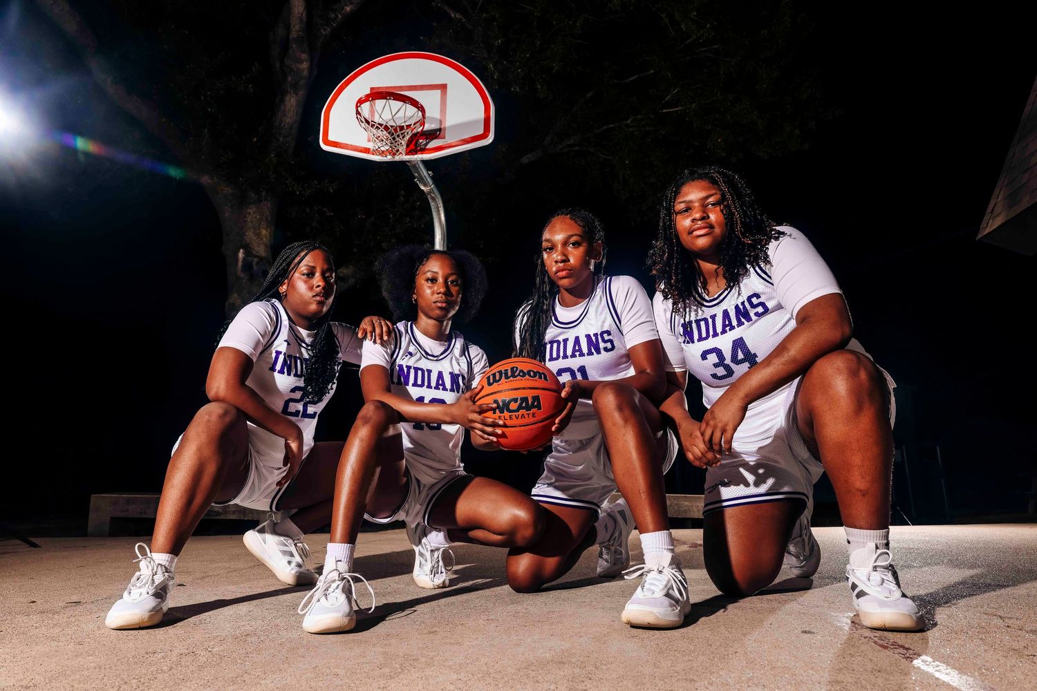 Basketball players in matching white uniforms pose together on the court at night under an illuminated hoop.