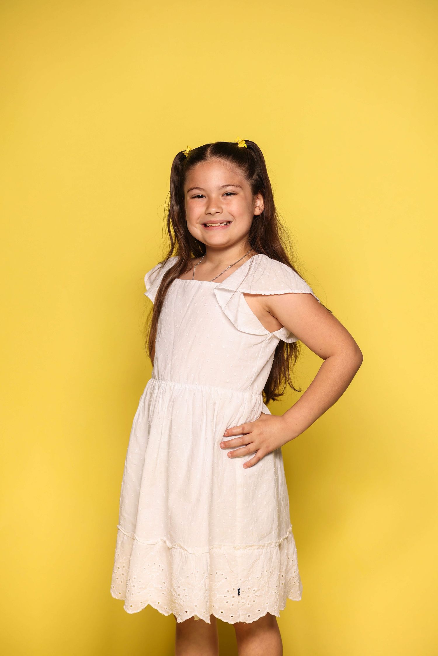Child in white eyelet dress poses playfully against bright yellow background in sequence of connected photos.