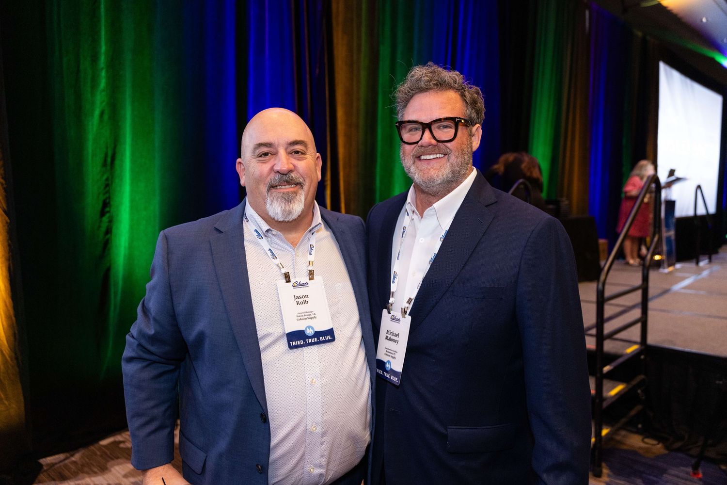 Two people in business attire smile together at a professional event with green lighting in the background.