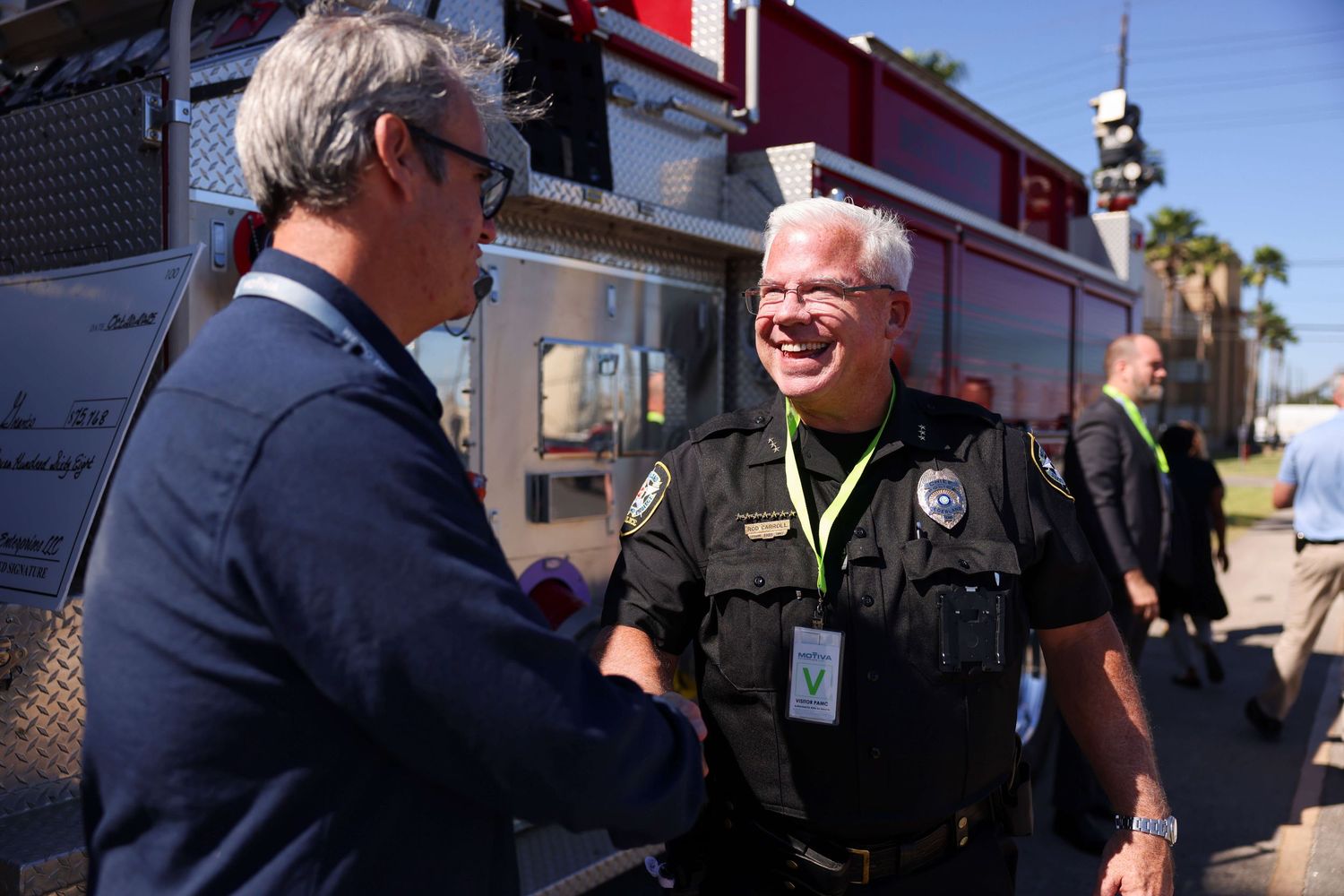 Two people shake hands and smile while standing in front of emergency response vehicles on a sunny day.