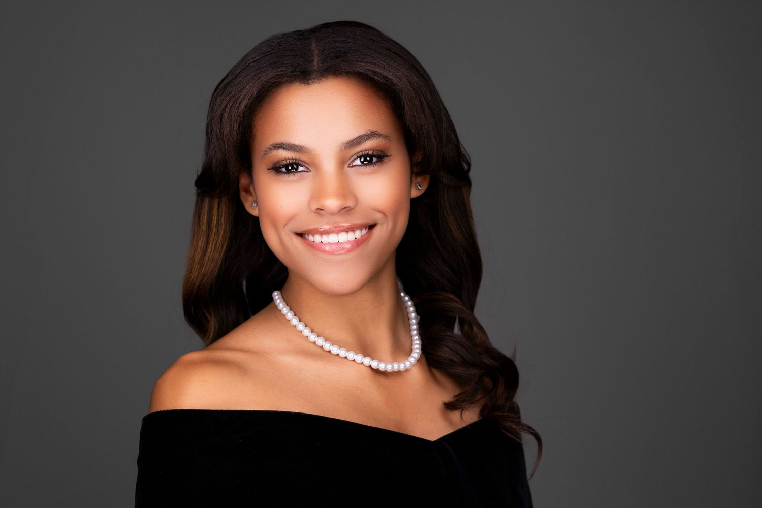 Professional portrait of someone wearing an off-shoulder black dress with pearl necklace and wavy dark hair against gray backdrop.