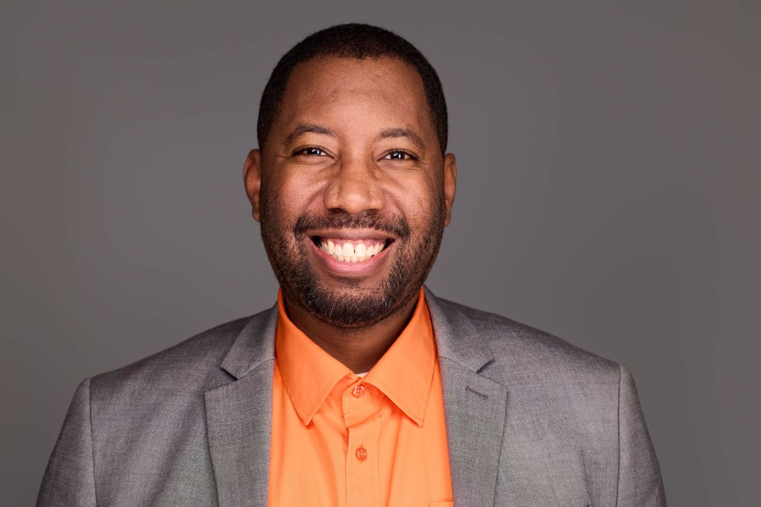 Professional portrait of someone wearing gray suit with orange polo against dark background.