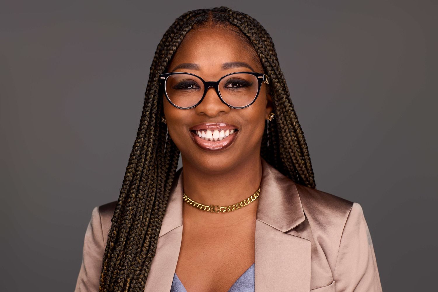 Professional portrait of someone in a beige blazer and glasses against a dark background.