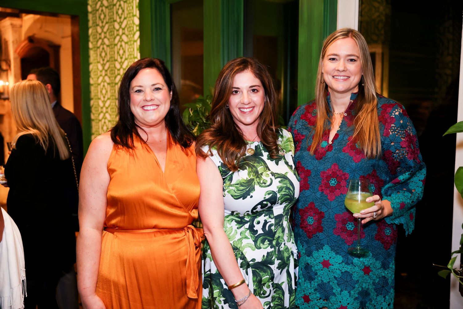 Three women in colorful dresses pose together at an elegant event with decorative wallpaper in the background.
