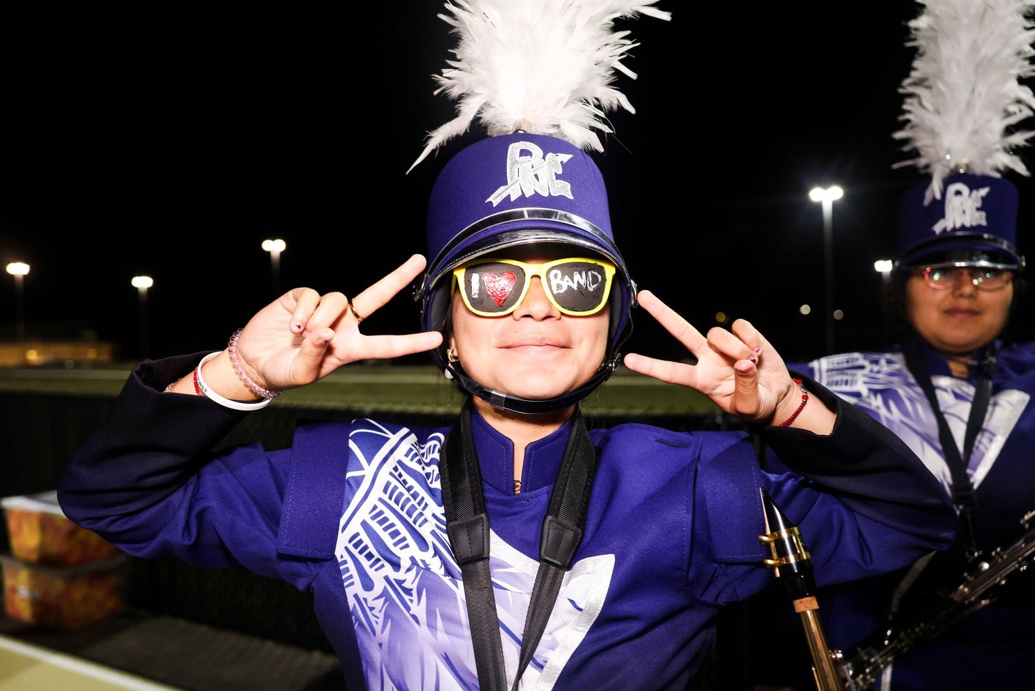 Band member in purple uniform with white feathered hat making peace sign gesture at night.