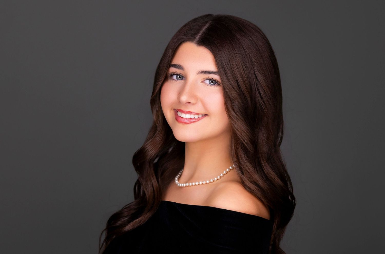 Studio portrait of someone wearing a black off-shoulder top and pearl necklace against a dark background.