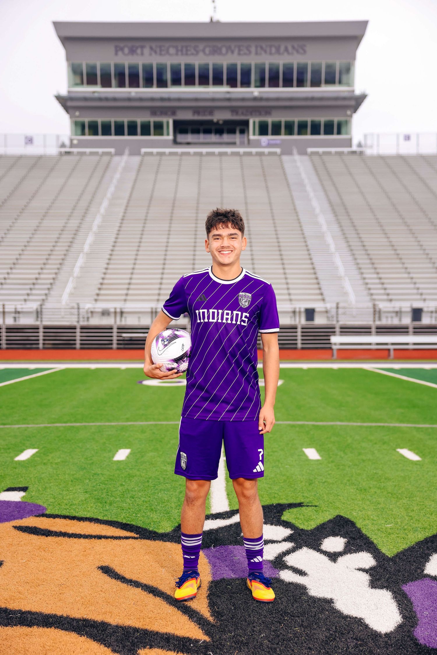 Soccer player in purple uniform stands on football field in front of stadium.