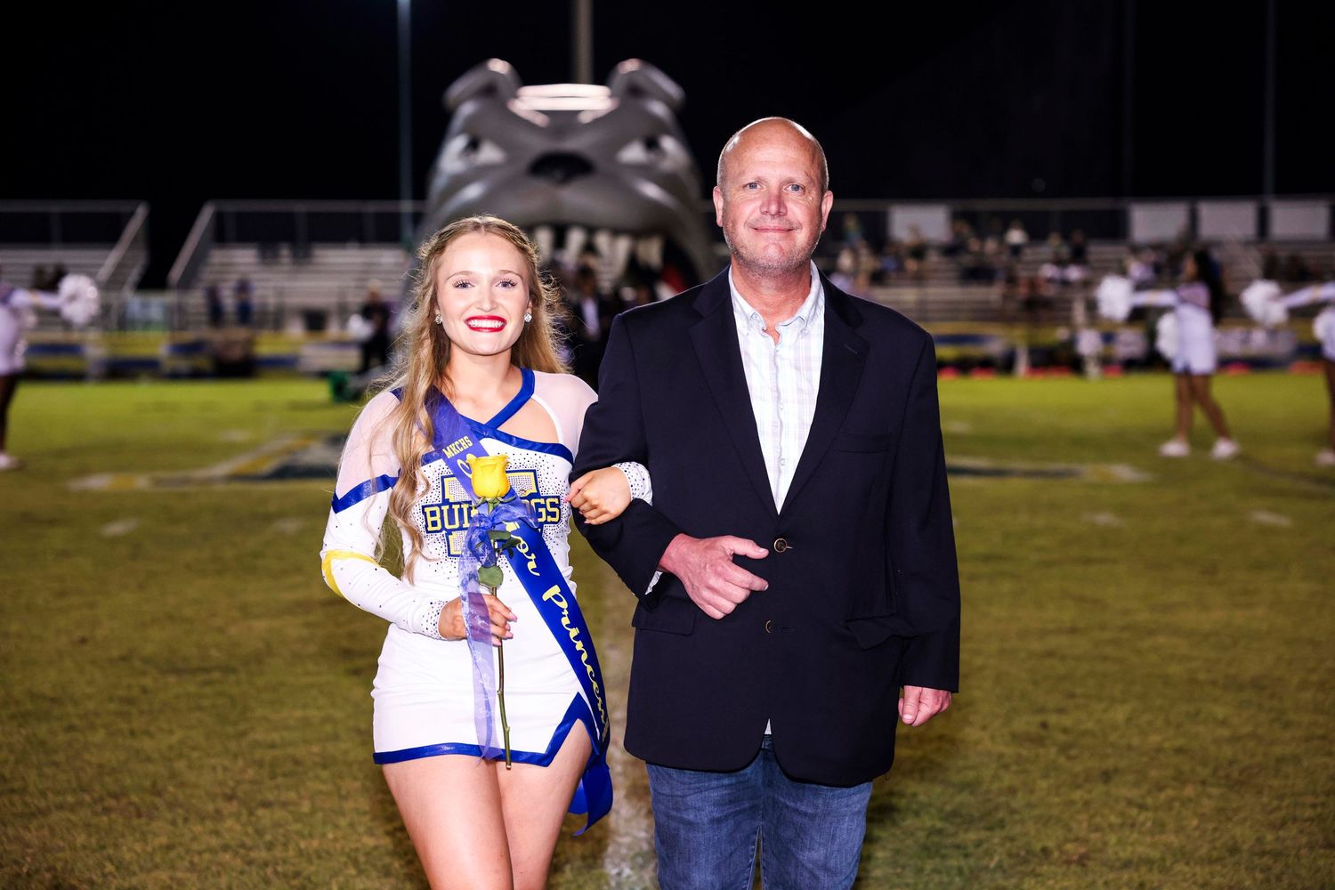 A cheerleader and adult stand together on a sports field during what appears to be a school event.