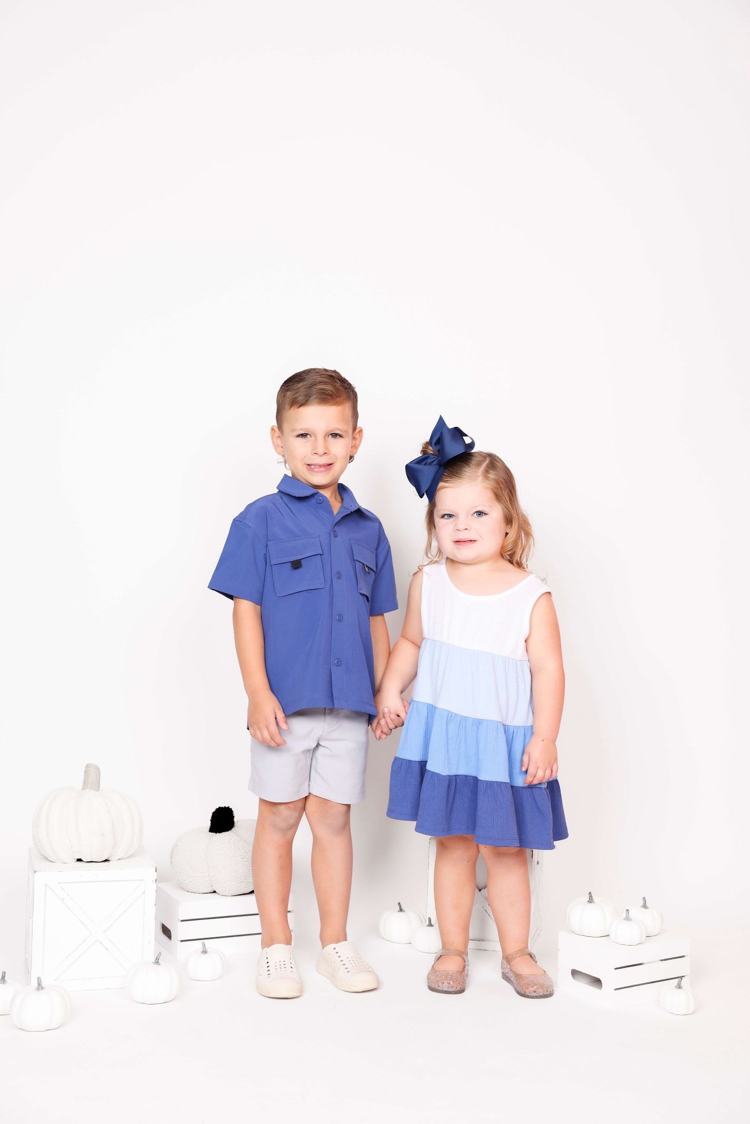 Two young children in coordinating blue and white outfits pose together on white blocks against a white backdrop.