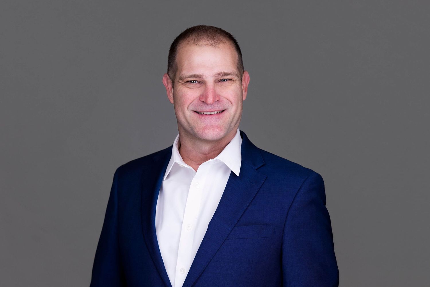 Business portrait of person wearing navy suit and white shirt against grey background.
