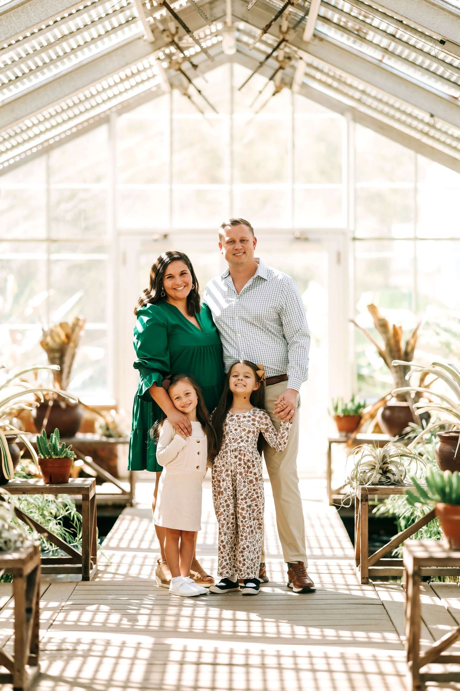 A family poses together in a bright sunlit greenhouse with potted plants.