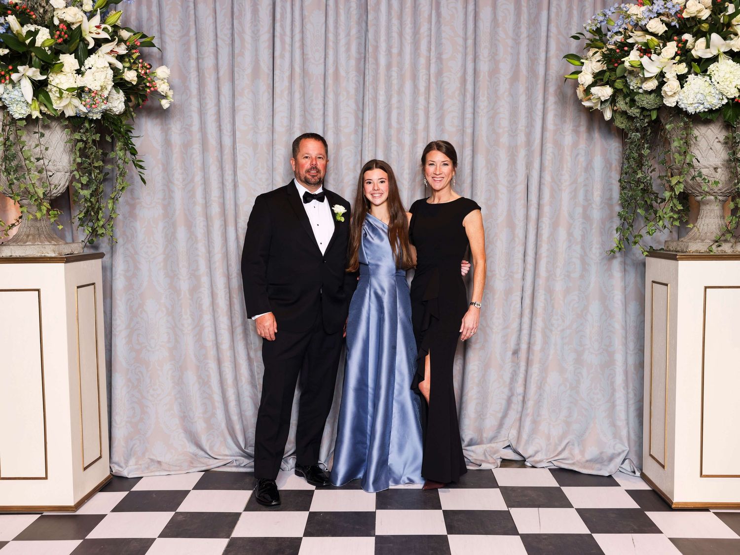 A group of wedding party members pose together in front of an elegant gray curtain backdrop with floral arrangements.