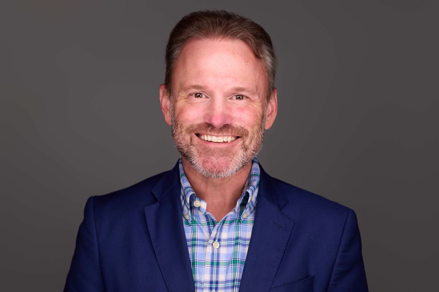 Professional headshot of person wearing blue suit and plaid shirt against dark background.