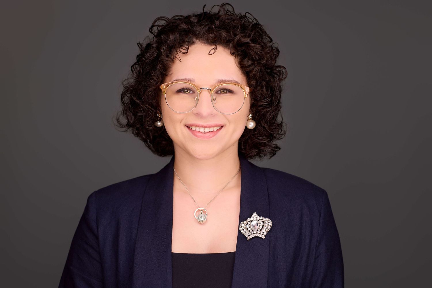 Professional headshot of someone with curly dark hair wearing a navy blazer against a gray background.