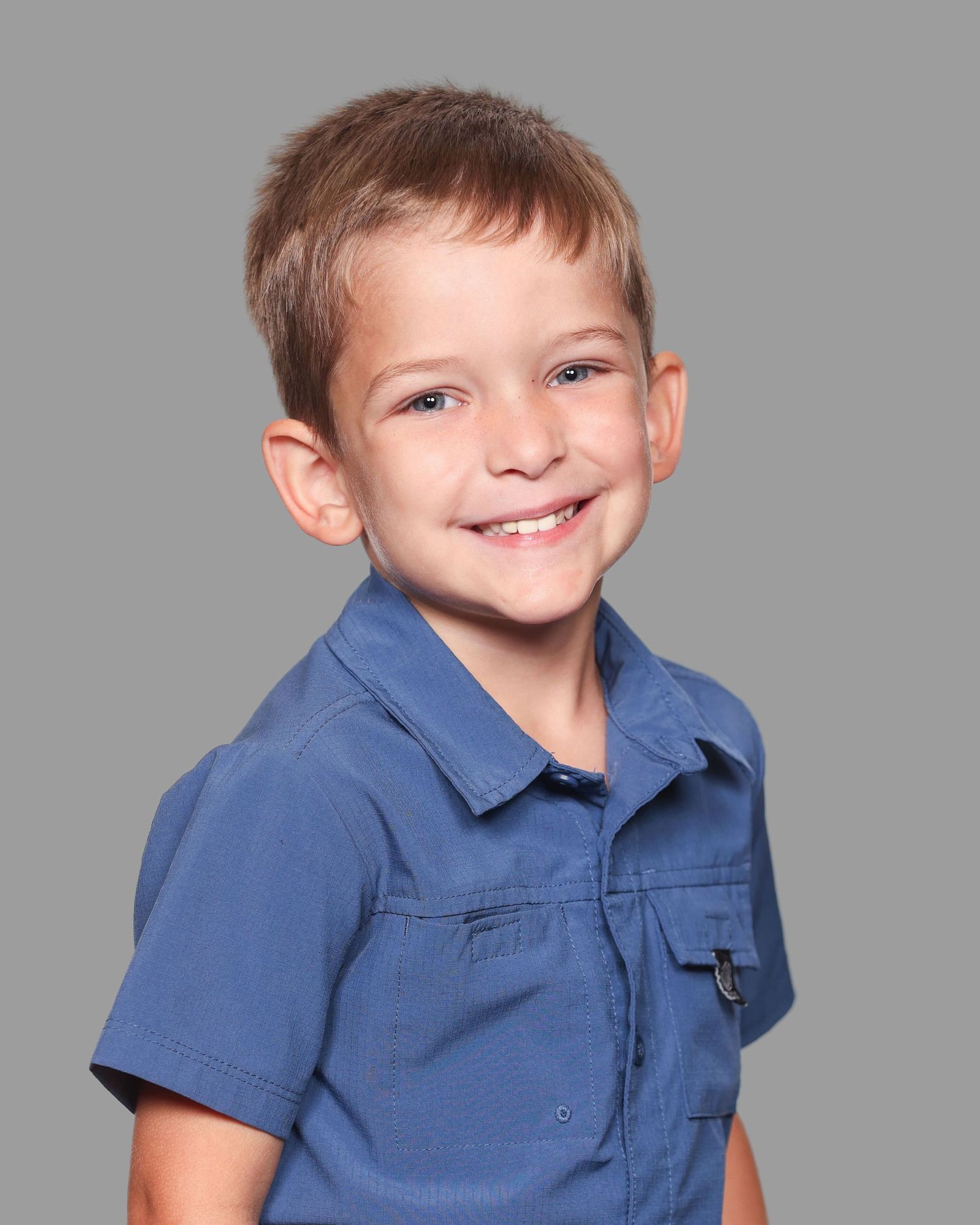 A young student wearing a blue collared shirt smiles warmly against a gray background in a school portrait.