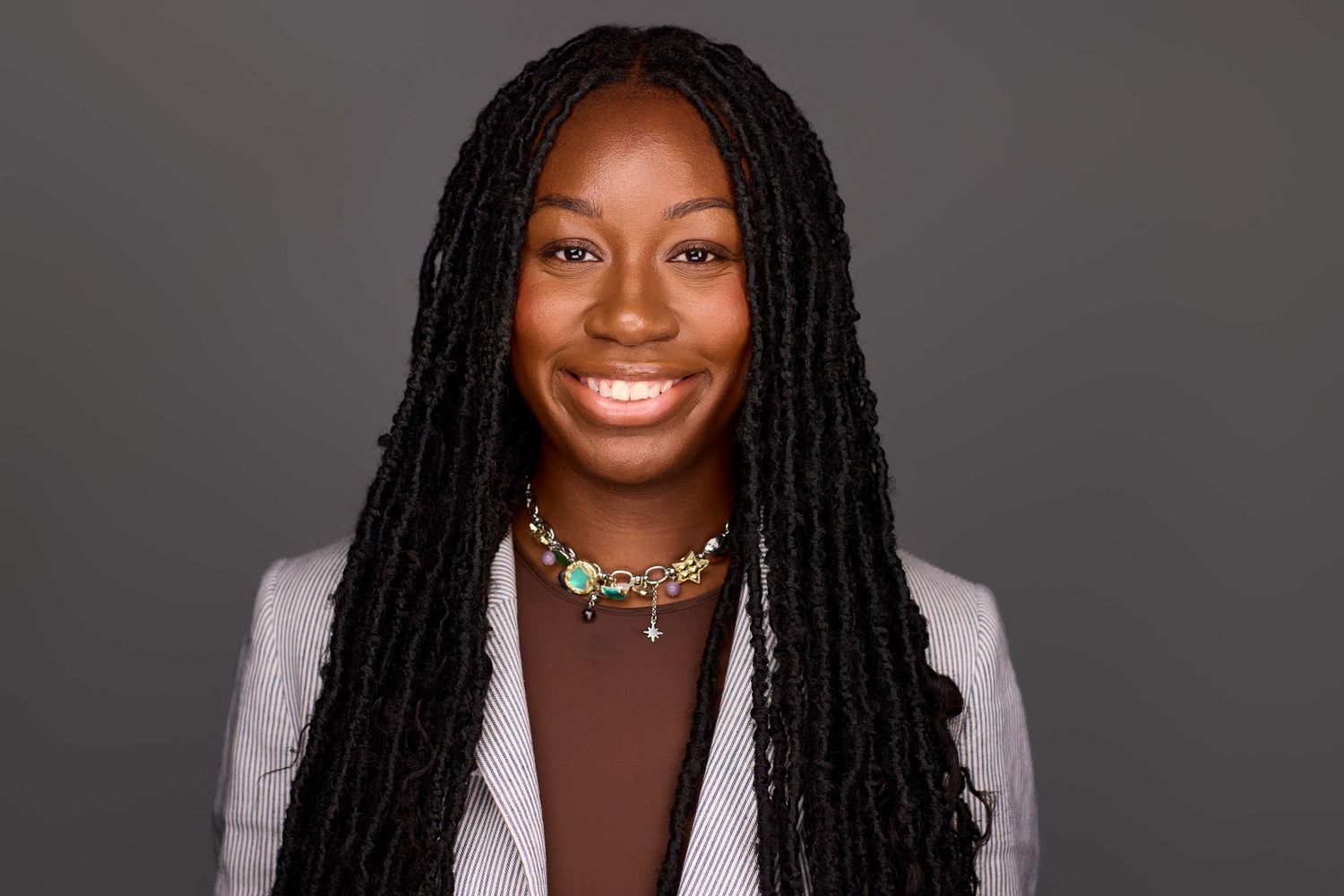 Professional portrait of person with braided hairstyle wearing white blazer against dark background.