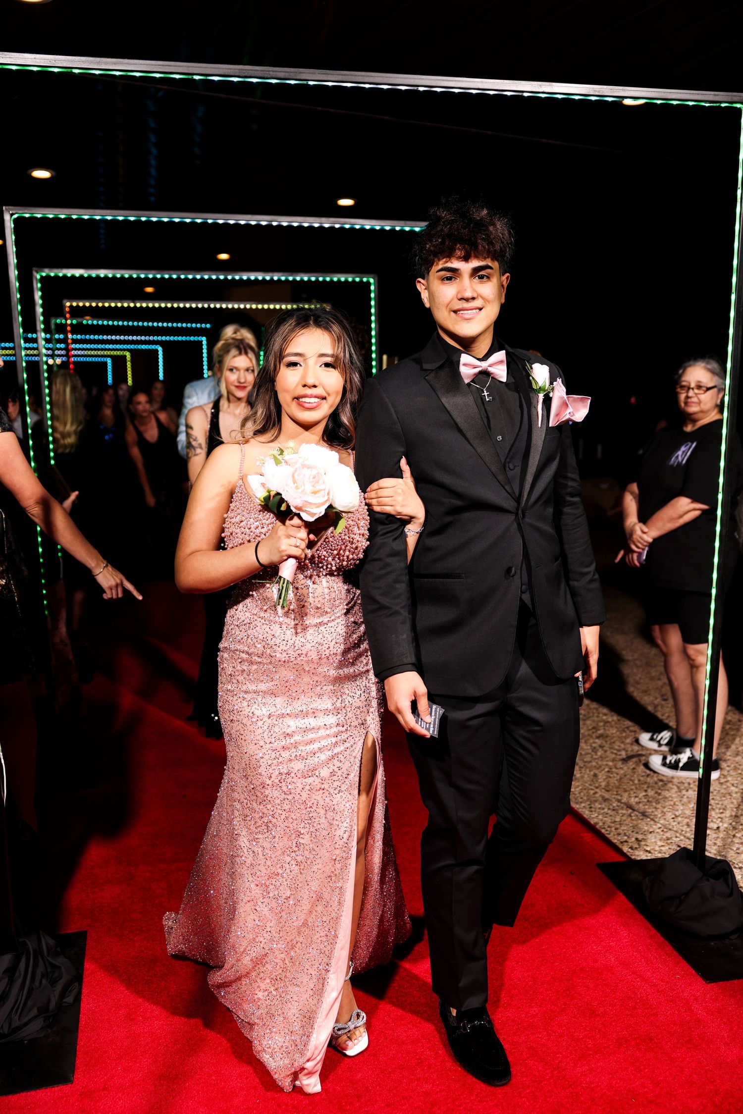 A couple walks together on a red carpet, with one person wearing a sparkly pink dress and the other in a black tuxedo.
